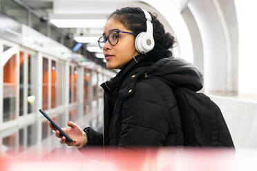 Side view of Colombian female in outerwear and eyeglasses scrolling social media on cellphone while listening to music in wireless headphones and waiting train on subway station - ADSF34612