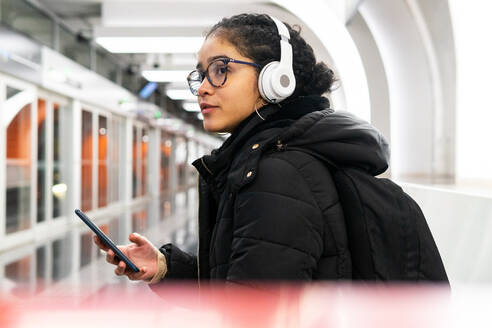 Seitenansicht einer kolumbianischen Frau in Oberbekleidung und mit Brille, die mit ihrem Handy in den sozialen Medien scrollt, während sie mit drahtlosen Kopfhörern Musik hört und in einer U-Bahn-Station auf den Zug wartet - ADSF34612