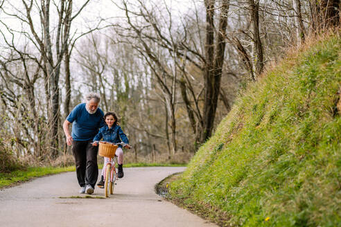 Fürsorglicher Großvater hilft aktiver Enkelin beim Fahrradfahren mit Korb auf der Straße in einer Landschaft mit grünen Bäumen an einem Sommertag - ADSF34613