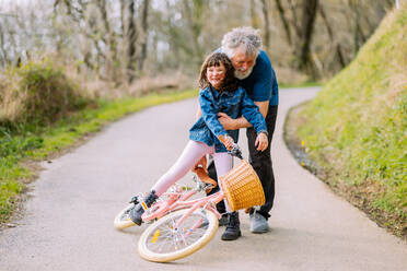 Full body of caring grandfather catching positive falling granddaughter from bicycle on path in park with trees on summer day - ADSF34620