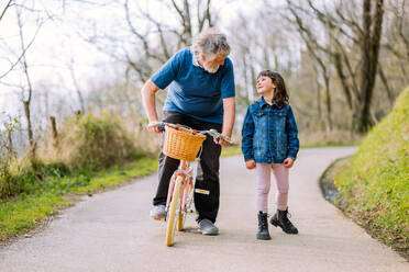 Full body of active grandfather riding bicycle of granddaughter near positive running girl on path in countryside with green trees - ADSF34621