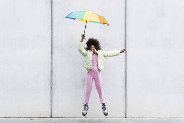 Glückliches Mädchen springt mit Regenschirm vor einer weißen Wand - PNAF03789