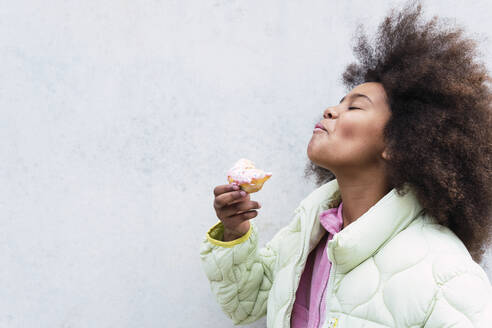 Girl with eyes closed enjoying eating doughnut in front of wall - PNAF03794
