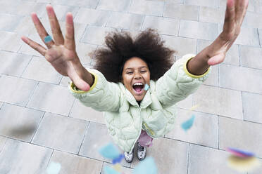 Cheerful girl with arms raised standing amidst confetti falling on footpath - PNAF03810