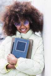 Smiling girl with curly hair holding book leaning on wall - PNAF03818