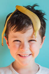 Headshot von fröhlichen Jungen mit Bananenschale auf dem Kopf Blick auf die Kamera mit Lächeln gegen blauen Hintergrund in Licht Studio - ADSF34628