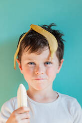 Headshot von fröhlichen Jungen mit Bananenschale auf dem Kopf und essen Banane, während in die Kamera schauen mit Lächeln gegen blauen Hintergrund in Licht Studio - ADSF34629