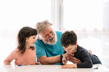 Fröhlicher bärtiger fürsorglicher Großvater sitzt in der Nähe von niedlichen Kindern, die mit Stiften auf Papier am Tisch in einem hellen Raum zu Hause schreiben - ADSF34635