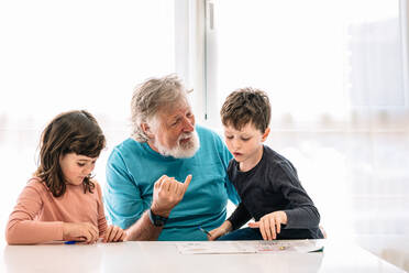 Positiver bärtiger fürsorglicher Großvater, der neben niedlichen Kindern sitzt, die mit Stiften auf Papier am Tisch in einem hellen Raum zu Hause schreiben - ADSF34636