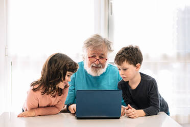 Bärtiger Großvater mit Brille und positiven Kindern, die ein interessantes Video auf einem Netbook ansehen, während sie am Tisch in einem hellen Raum zu Hause sitzen - ADSF34637