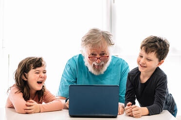 Bärtiger Großvater mit Brille, der die Zunge herausstreckt, mit fröhlichen Kindern, die ein interessantes Video auf einem Netbook ansehen, während sie am Tisch in einem hellen Raum zu Hause sitzen - ADSF34638