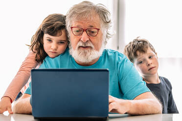 Bärtiger Großvater mit Brille und Kindern, die ein interessantes Video auf einem Netbook ansehen, während sie am Tisch in einem hellen Raum zu Hause sitzen - ADSF34639