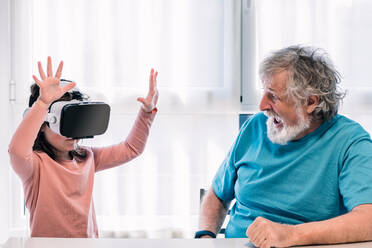 Niedliches Mädchen mit moderner VR-Brille, das mit erhobenen Armen den Cyberspace erkundet, während es neben seinem bärtigen Großvater in einem hellen Raum zu Hause sitzt - ADSF34655
