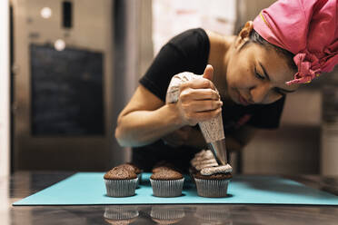 Hispanic female baker in headscarf using pastry bag to squeeze cream on top of chocolate cupcakes while working in bakehouse - ADSF34663