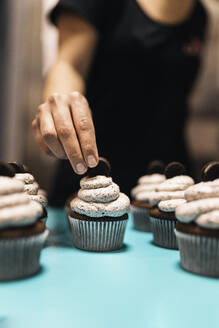 Anonyme Frau, die einen winzigen Keks auf ein frisches Törtchen vor einem unscharfen Hintergrund legt, während sie in einer Bäckerei arbeitet - ADSF34664
