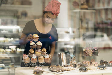 Hispanische Frau mit Kopftuch arrangiert Desserts auf einem Tisch in der Nähe des Fensters, während sie tagsüber in einer Bäckerei arbeitet - ADSF34666