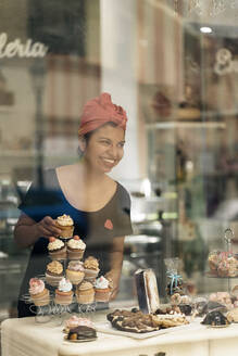 Hispanische Frau mit Kopftuch arrangiert Desserts auf einem Tisch in der Nähe des Fensters, während sie tagsüber in einer Bäckerei arbeitet - ADSF34667