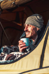 Male hiker in warm clothes lying on blanket in camping tent and drinking cup of coffee in morning and admiring nature at dawn - ADSF34680