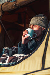 Male hiker in warm clothes lying on blanket in camping tent and drinking cup of coffee in morning and admiring nature at dawn - ADSF34681