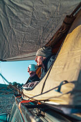 Männlicher Wanderer in warmer Kleidung liegt auf einer Decke im Campingzelt und trinkt eine Tasse Kaffee am Morgen und bewundert die Natur in der Morgendämmerung - ADSF34682