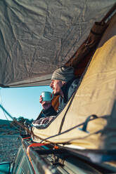 Male hiker in warm clothes lying on blanket in camping tent and drinking cup of coffee in morning and admiring nature at dawn - ADSF34682