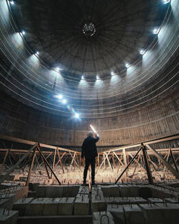 Unbekannte Person mit heller Lampe, die auf Betonblöcken steht und eine große Kuppel in einem desolaten Kraftwerk untersucht - ADSF34685