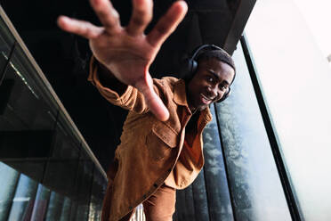 From above of positive African American male looking at camera and reaching out hand while listening to music in headphones on street - ADSF34693