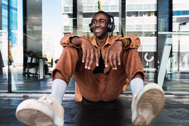 Stylish African American male listening to music in wireless headphones and looking away while sitting near glass wall in city - ADSF34694