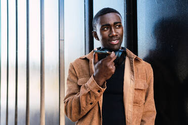 Emotionless African American male with black wireless headphones recording voice message on smartphone while standing near metal wall in city - ADSF34696