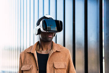 Afroamerikanischer Mann mit VR-Brille, der den Cyberspace erkundet, während er auf der Straße in der Nähe eines modernen Gebäudes mit Metallwand in einer Stadt steht - ADSF34697