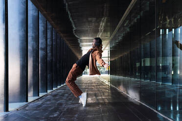 Ganzkörper-Seitenansicht eines aktiven afroamerikanischen Mannes, der auf Zehenspitzen steht und sich nach hinten beugt, während er in der Nähe eines modernen Gebäudes Musik hört - ADSF34698