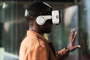 Positive African American male in modern VR goggles standing near glass wall while exploring virtual reality on street of city - ADSF34699