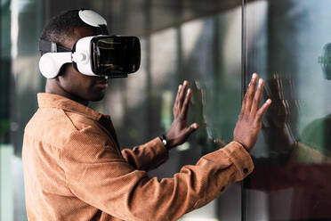 Positive African American male in modern VR goggles standing near glass wall while exploring virtual reality on street of city - ADSF34700