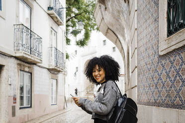 Smiling woman holding mobile phone standing on footpath - DCRF01133