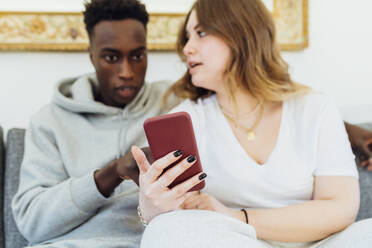 Multiracial couple with smart phone sitting on sofa at home - MEUF05319