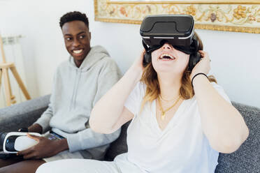 Glückliche Frau, die einen Virtual-Reality-Simulator trägt und mit ihrem Freund auf dem Sofa zu Hause sitzt - MEUF05328