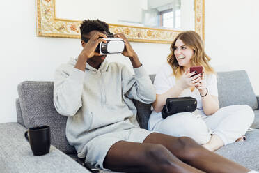 Man wearing virtual reality simulator sitting by girlfriend on sofa at home - MEUF05329