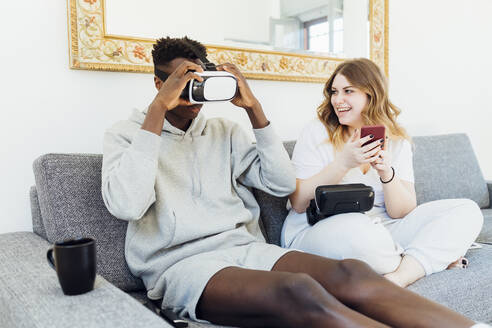 Mann mit Virtual-Reality-Simulator sitzt mit seiner Freundin auf dem Sofa zu Hause - MEUF05329