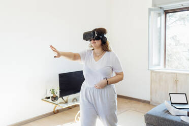 Young woman wearing virtual reality simulator gesturing in living room at home - MEUF05332