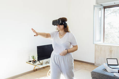 Junge Frau trägt einen Virtual-Reality-Simulator und gestikuliert im heimischen Wohnzimmer - MEUF05332