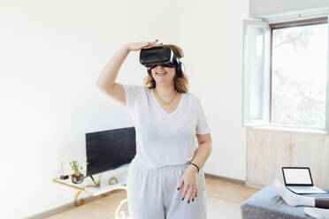 Glückliche Frau, die einen Virtual-Reality-Simulator trägt, steht im Wohnzimmer zu Hause - MEUF05333