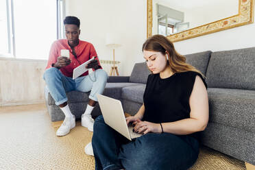 Multiracial couple using laptop and mobile phone in living room at home - MEUF05338