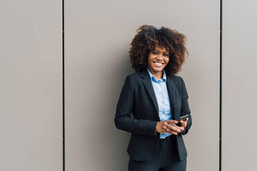 Happy Afro businesswoman holding mobile phone standing in front of wall - MEUF05381