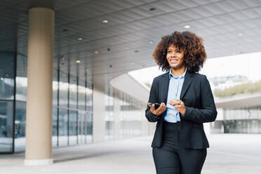 Smiling businesswoman holding smart phone walking outside office building - MEUF05388