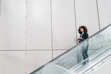 Afro-Geschäftsfrau mit Tablet-PC, die auf einer Rolltreppe nach unten fährt - MEUF05395