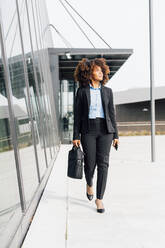 Businesswoman holding laptop bag walking by glass wall - MEUF05401