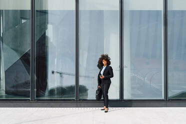 Businesswoman holding laptop bag and mobile phone standing in front of glass wall - MEUF05403