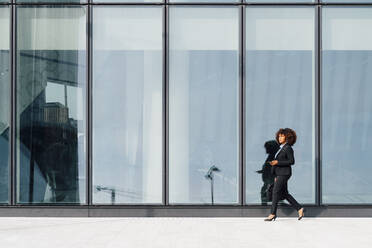 Businesswoman holding laptop bag and mobile phone walking by glass wall - MEUF05404