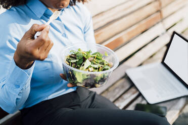 Businesswoman eating salad sitting on bench - MEUF05436