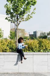 Businesswoman holding salad lunch box sitting legs crossed on concrete wall - MEUF05440